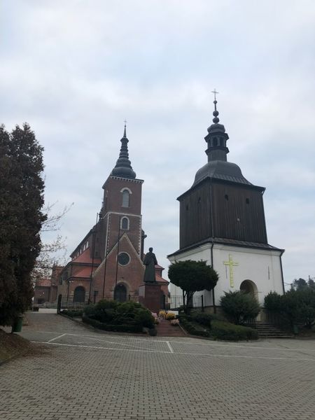 Ceglany kościół parafialny w Niegowici z wysoką wieżą, obok biała wieża z drewnianą górną częścią, przed nimi pomnik i brukowany plac.