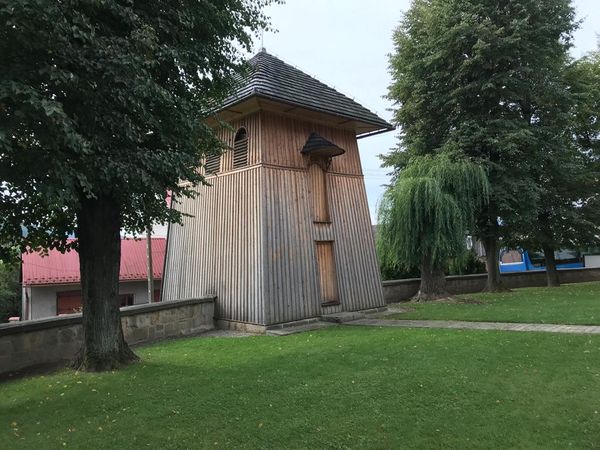 Drewniana dzwonnica w Raciechowicach o zwężającej się ku górze bryle, na trawniku przy kamiennym murze, wśród drzew i wierzby płaczącej.