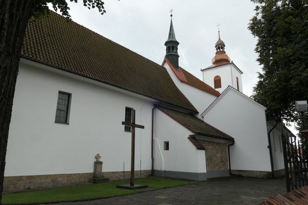 Biały kościół w Gdowie z boku: długi dach z ciemnej dachówki, dwie wieżyczki z hełmami, przed ścianą drewniany krzyż i kamienny nagrobek.