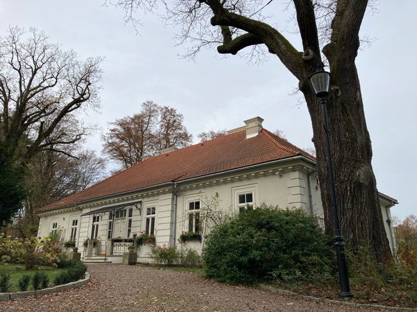 Biały parterowy dwór w Hucisku z dachem z czerwonej dachówki i kwiatami pod oknami, przed nim żwirowy podjazd, stare drzewo i latarnia.