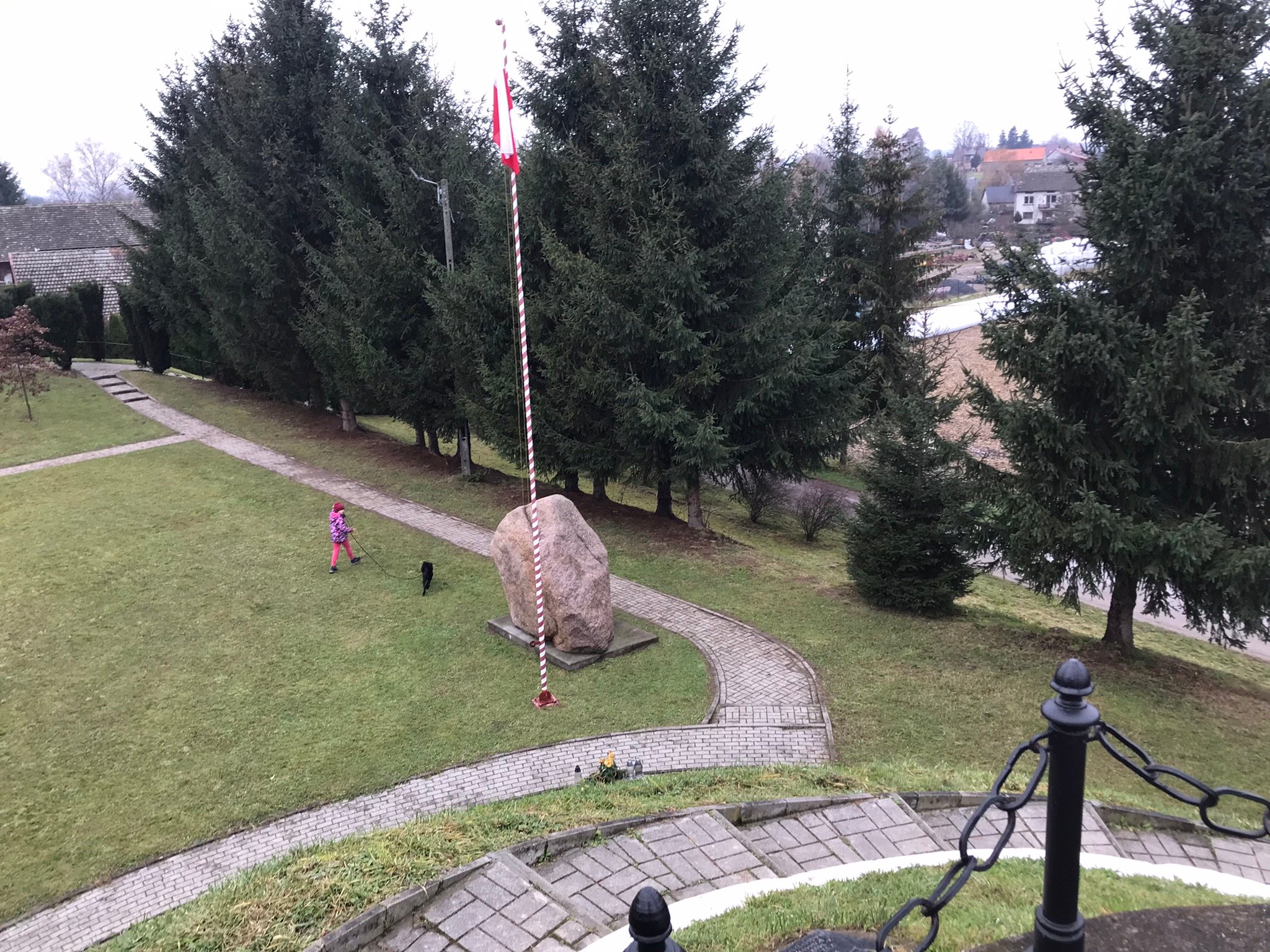 Widok ze szczytu kopca na duży głaz i maszt z flagą przy alejce; po trawniku dziecko prowadzi czarnego psa, w tle świerki.