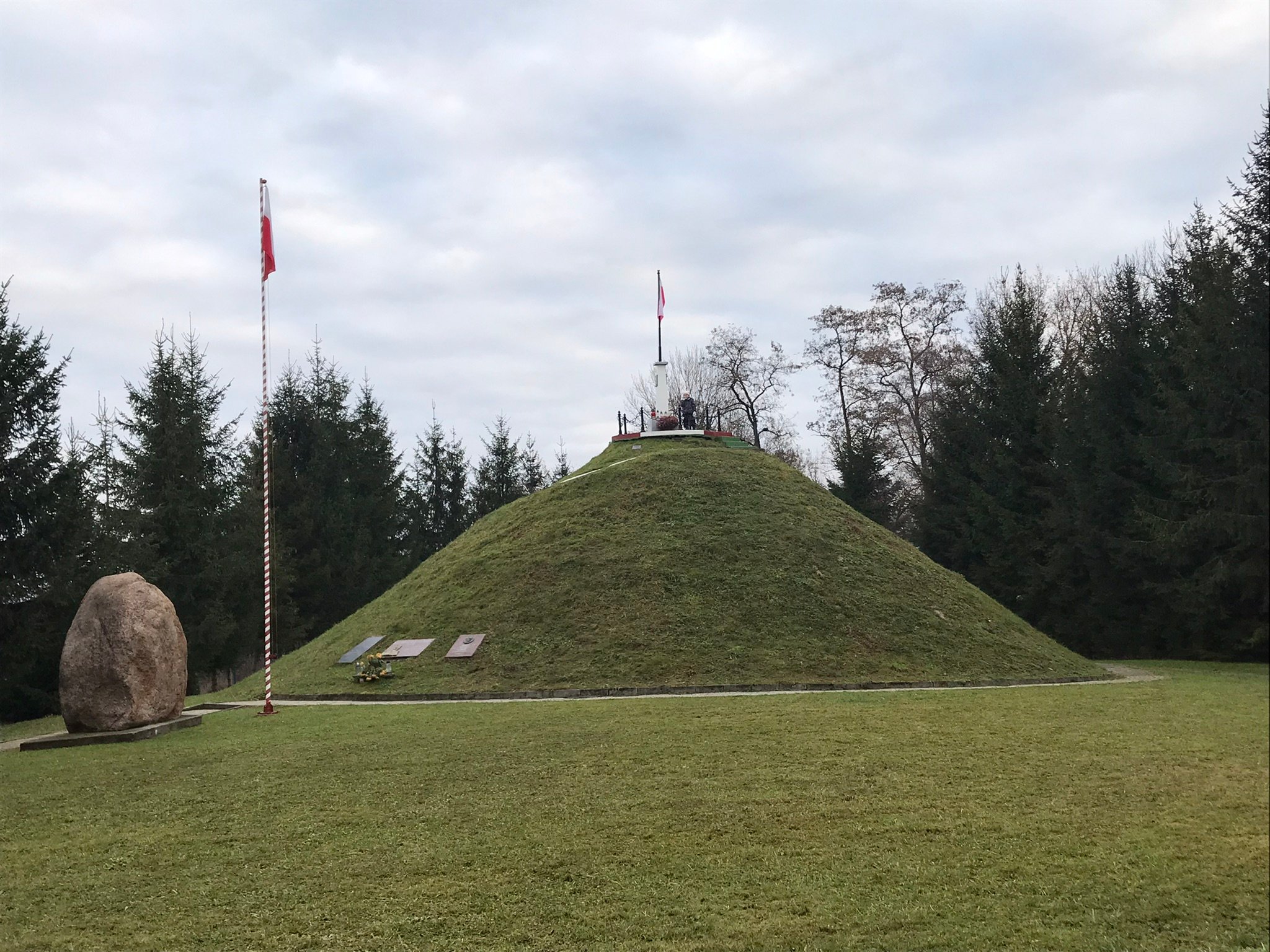Trawiasty kopiec gen. Jana Henryka Dąbrowskiego w Pierzchowie z flagą na szczycie, obok wysoki maszt z flagą, duży głaz i świerki.