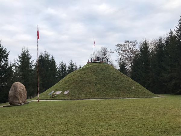 Trawiasty kopiec gen. Jana Henryka Dąbrowskiego w Pierzchowie z flagą na szczycie, obok wysoki maszt z flagą, duży głaz i świerki.