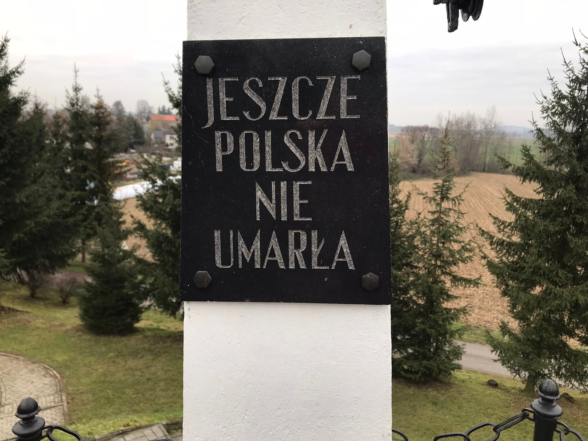 Czarna tablica z napisem „Jeszcze Polska nie umarła” na białym słupie na szczycie kopca, w tle świerki, pola i domy.
